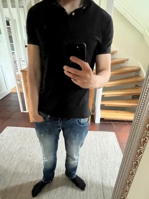 Svart ralph lauren pikétröja med krage - Klassisk svart pikétröja med krage och knappar framtill. Tröjan har korta ärmar och en slim passform som sitter snyggt på kroppen. Perfekt till jeans eller chinos för en clean look.