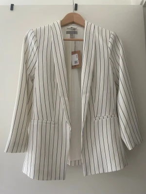 Vit kavaj med svarta ränder H&M - Snygg vit kavaj från H&M med tunna svarta vertikala ränder. Rak passform, två fickor framtill och utan knäppning för en clean look. Perfekt till både jeans och kostymbyxor. Materialet känns lätt och stilrent.