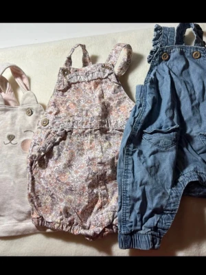 Blommig byxdress & blå hängselbyxa - Två söta plagg: en blommig byxdress i pastellfärger med volangdetaljer och knappar, samt ett par blå hängselbyxor i mjukt denimliknande material med fickor och resår vid benslut. Perfekt för en lekfull och trendig stil.