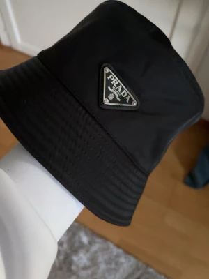 Svart Prada Hatt Bucket Hat - Snygg svart bucket hat från Prada med den ikoniska trekantiga logotypen framtill. Hatten är tillverkad i ett glansigt syntetmaterial som ger en modern och clean look. Perfekt accessoar för att lyfta din outfit.