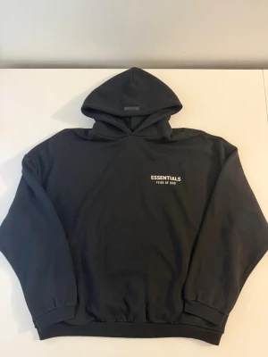 Svart Essentials Fear of God hoodie - Säljer en svart hoodie från Essentials Fear of God med vit logga tryckt på bröstet. Hoodien har en klassisk passform med huva och ribbade muddar vid ärmar och midja. Perfekt för en clean och stilren streetwear-look.