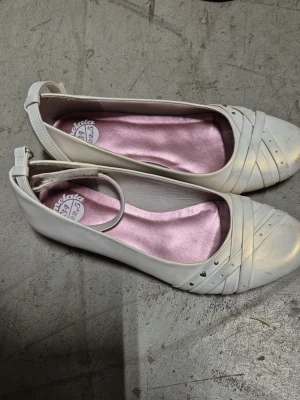 Ballerinaskor - Snygga beige guldiga ballerinaskor från Marmelata med dekorativa remmar och små silverfärgade nitar på tån. Skorna har en smal rem runt ankeln och rosa innersula i satinliknande material. Perfekt för dig som gillar stilrena och feminina skor.