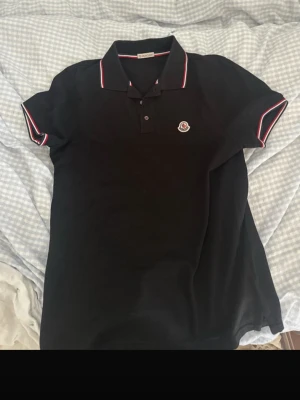 Svart Moncler pikétröja med röd/vit kant - Klassisk svart pikétröja från Moncler med korta ärmar och vit/röd kant på krage och ärmslut. Tröjan har knappslå framtill och Moncler-logga på bröstet. Perfekt till en stilren och sportig look.