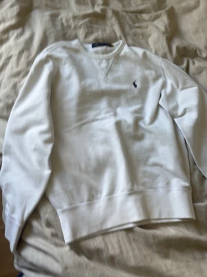 Vit sweatshirt från Polo Ralph Lauren - Klassisk vit sweatshirt från Polo Ralph Lauren med rund halsringning och diskret broderad logga på bröstet. Tröjan har ribbade muddar vid ärmslut och nederkant, perfekt för en clean och stilren look.