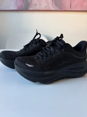 Hoka womens bondi 9 black - Säljer ett par helsvarta sneakers från Hoka med riktigt chunky och hög sula. Skorna har mesh-ovandel för bra andningsförmåga och klassisk snörning. Perfekta för dig som gillar sportig stil och vill ha extra komfort i steget.