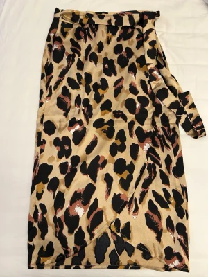 Leopardmönstrad omlottkjol beige från Boohoo - Omlottkjol i beige med leopardmönster i svart, brunt och vitt från Boohoo i storlek 34. Kjolen har knytband i midjan och är i ett mjukt, glansigt material. 