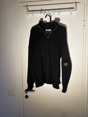 Stone Island Zip - Säljer en svart stickad ziptröja från Stone Island med ikonisk patch på vänster ärm. Tröjan har hög krage, dragkedja framtill och ribbade muddar. Perfekt för dig som gillar streetwear och vill ha något stilrent men ändå med attityd.