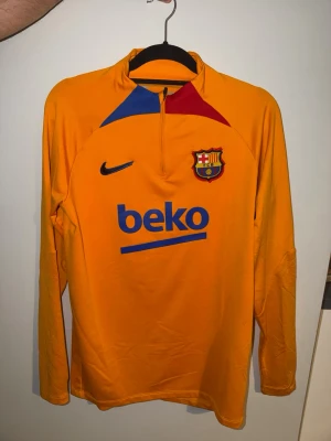 Barcelona träningströja Nike  -  - Gul långärmad träningströja från Nike med FC Barcelona-logga och Beko-tryck på både fram- och baksida. Tröjan har halv dragkedja, blå och röd detalj vid halsen och är tillverkad i ett lätt funktionsmaterial som passar perfekt till träning. 