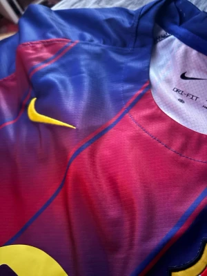 Nike Barcelona matchtröja blå/röd - Säljer en officiell Barcelona matchtröja från Nike i blått och rött med gula detaljer. Tröjan har tryckta märken och Champions League-patch på ärmen. Tillverkad i lätt och ventilerande polyester med Dri-FIT-teknologi, perfekt för fotboll eller träning.