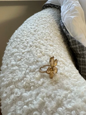 Guldfärgad blomring i metall - Säljer en statementring i guldfärgad metall med en stor blomma som motiv. Ringen har fem rundade blomblad och en blank finish som fångar ljuset snyggt. Perfekt för dig som vill ha en unik accessoar med blommig design.