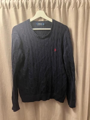 Mörkblå kabelstickad tröja Polo Ralph Lauren - Kabelstickad mörkblå tröja från Polo Ralph Lauren med rund halsringning och röd broderad logga på bröstet. Tröjan har långa ärmar och ribbade muddar vid ärmslut och nederkant. Perfekt för lager på lager och klassisk stil. Det är ett litet hål på axeln, kolla bilden.