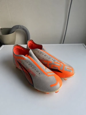 Puma Ultra grå och orange fotbollsskor - Snygga fotbollsskor från Puma i vitt och orange med svart logga på sidan. Skorna har en strömlinjeformad design, låg vikt och ovandel i syntetmaterial. Sulan är vit med orange dobbar för bra grepp på planen. Perfekta för dig som vill ha snabbhet och stil på matchen. Snören kommer med om man vill ha. PRIS KAN PRUTAS.