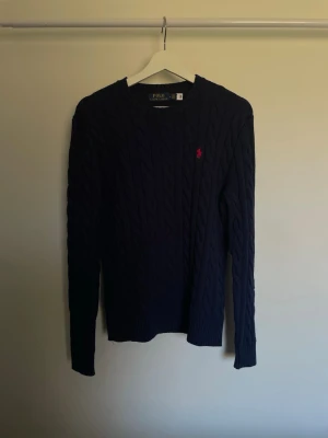 Mörkblå kabelstickad tröja Polo Ralph Lauren - Mörkblå kabelstickad tröja från Polo Ralph Lauren med rund halsringning och lång ärm. Tröjan har det klassiska röda broderade logotypen på bröstet och ribbstickade muddar vid ärmslut och nederkant. Perfekt för en stilren och tidlös look.
