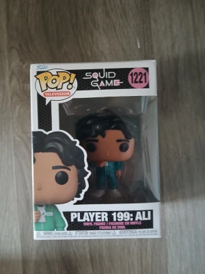 Funko Pop Squid Game Ali #1221 - Cool Funko Pop-figur från Squid Game, Player 199: Ali. Figuren är gjord i vinyl och kommer i originalförpackning med fönster. Perfekt för samlare eller som dekoration. Färgerna är mestadels svart, brun och turkos.
