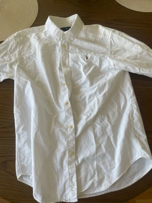 Vit skjorta Polo Ralph Lauren - Klassisk vit skjorta från Polo Ralph Lauren med button-down krage och broderad logga på bröstet. Skjortan har lång ärm, knäppning framtill och är tillverkad i mjuk bomull. Perfekt till en stilren och clean look.
