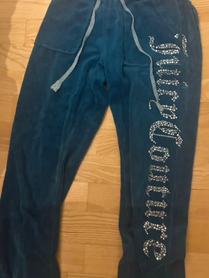 Blå velourbyxor Juicy Couture - Mörkblå mjukisbyxor i velour från Juicy Couture med stora strasstenar som bildar loggan längs ena benet. Byxorna har dragsko i midjan och två fickor med lock och knapp bak. Perfekta för en chill och trendig look.