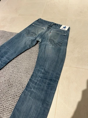 Blå raka jeans från Levi's - Säljer ett par klassiska blå jeans från Levi's med raka ben och femficksmodell. Jeansen har en snygg tvättad look och tydliga sömmar på bakfickorna. Tillverkade i slitstark denim, perfekta för en avslappnad stil.