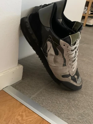 Valentino camo sneakers i grått - Säljer ett par snygga Valentino sneakers med grå, svart och beige camo-mönster. Skorna har låga skaft, svarta sulor och vita snören. Materialmix av mocka och textil ger en cool och unik look. Perfekta för dig som vill sticka ut med stil.