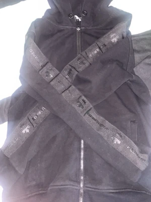 Svart hoodie från Dsquared2 - Säljer en svart hoodie från Dsquared2 med dragkedja framtill och broderad logga på bröstet. Hoodien har två fickor och en klassisk huva. Materialet känns mjukt och bekvämt, perfekt för en avslappnad streetwear-look.