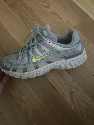 Nike grå sneakers med pastellfärgardetaljer - Nike sneakers i grått med inslag av pastellrosa och lila detaljer samt en gul swoosh. Skorna har en chunky vit sula och mesh- och syntetmaterial i ovandelen. Snygg sportig look med coola färgskiftningar och klassisk Nike-logga på sidan. Limited edition