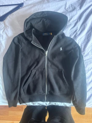 Svart hoodie från Polo Ralph Lauren - Klassisk svart hoodie från Polo Ralph Lauren med dragkedja framtill och huva. Liten vit broderad logga på bröstet och två fickor fram. Tillverkad i mjukt material som känns skönt mot huden. Perfekt för en avslappnad och stilren look. Har lite katthår på men kan ta bort innan skick! KOM DM FÖR MER INFO!