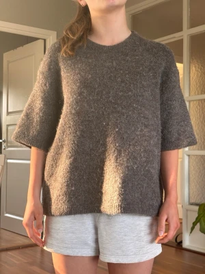 Brun stickad tröja med kort ärm - I premiumkvalitet i 14% ull. Mysig brun stickad tröja med rund hals och halvlånga, vida ärmar. Tröjan har en avslappnad passform och är gjord i ett mjukt, luddigt material som känns skönt mot huden. Perfekt för lager på lager och enkel att styla till olika outfits.