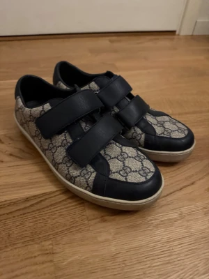 Gucci sneakers med monogram och kardborre - Snygga sneakers från Gucci med klassiskt GG-monogram i beige och mörkblå detaljer. Skorna har två breda kardborreband och vit sula. Perfekta för dig som vill ha en stilren och lyxig look med streetkänsla. Har även låda, påsen och ett äkthetsintyg från vestiere. 