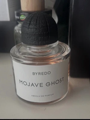 Byredo Mojave Ghost parfym ca 55 ml kvar - Byredo Mojave Ghost Absolu de Parfum i en rund, genomskinlig glasflaska med svart räfflad kork. Parfymen har en varm bärnstensfärgad vätska och minimalistisk vit etikett med svart text. En stilren och modern doftflaska som passar perfekt på parfymhyllan. 