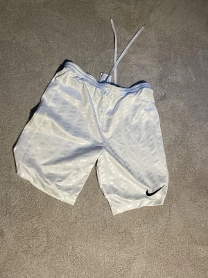 Vita Nike Dri-Fit shorts  - Snygga vita sportshorts från Nike med Dri-Fit-teknologi och diskret mönster i tyget. Shortsen har elastisk midja med snörning och svart Nike-logga på benet. Perfekta för träning eller sportiga dagar.