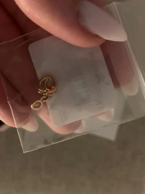 Guldigt Pandora halsbandhänge hjärta  - Säljer ett guldigt halsbandhänge från Pandora med hjärtdesign och en liten röd sten. Hänget har en elegant och stilren look, perfekt att fästa på ditt favoritarmband från Pandora. Kommer i originalask.