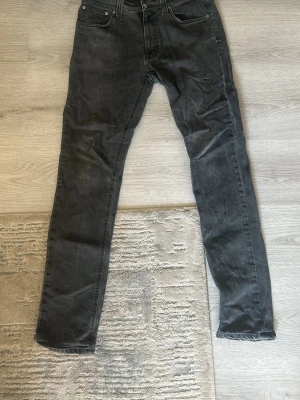 Svarta skinny jeans från Nudie Jeans - Snygga svarta skinny jeans från Nudie Jeans med klassisk femficksdesign och silverfärgade nitar. Jeansen har en något tvättad look och smal passform som sitter tajt längs benen. Perfekta för en stilren och modern outfit.