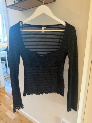 Svart ribbad långärmad topp - Trendig svart ribbad topp med lång ärm och lätt croppad passform. Toppen har en snygg v-ringning och diskret rynkdetalj framtill. Perfekt att styla med både jeans och kjol för en cool look.