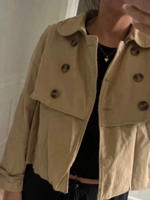 Kort beige trenchcoat med stora knappar - Snygg kort beige trenchcoat med dubbelknäppning och stora bruna knappar. Jätte fin!!🩷