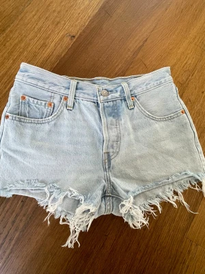 Ljusblå Levi's jeansshorts med fransar W25 - Säljer ett par ljusblå jeansshorts från Levi's med hög midja och rå franskant. Klassisk femficksdesign, knapp och dragkedja framtill samt Levi's patch och röd etikett bak. Perfekta för sommarens alla äventyr.