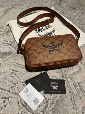 MCM Himmel crossbody - Snygg brun axelväska från MCM i klassiskt monogrammönster med svart logotyp och guldiga detaljer. Väskan har justerbar axelrem och är tillverkad i tåligt läder. Perfekt storlek för det viktigaste och levereras med dustbag och äkthetskort.