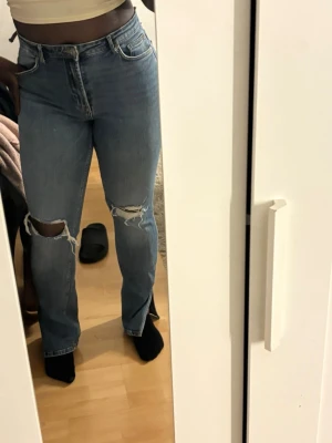 Blå slitna jeans med hål på knäna - Säljer ett par blå jeans med raka ben och coola slitningar på båda knäna. Jeansen har klassisk femficksdesign, hög midja och är gjorda i denim. 