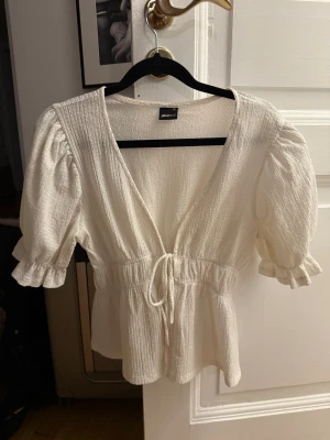 Vit blus med knyt från Gina Tricot  - Söt vit blus från Gina Tricot med v-ringning, puffärmar och knyt i midjan. Tillverkad i ett lätt, strukturerat material som ger en luftig känsla. Perfekt för en trendig och avslappnad look.