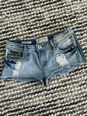 Blå denim shorts med nitar - Säljer ett par blå denim shorts från Parisian Collection. Shortsen har slitna detaljer, nitar längs fickorna och ett broderat mönster i turkost, svart och vitt på ena fickan. Klassisk femficksmodell med råa kanter och låg midja. Storlek 38 men passar för s-xs