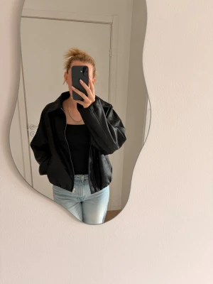 Oversized svart skinnjacka/bomberjacka - Säljer en oversized svart skinnjacka med bred krage och dragkedja framtill. Jackan har en avslappnad passform och går in vid midjan. Modellen kiknar en bomberjacka. Aldrig använd och prislapp kvar. Storlek XS. Nypris 419kr. 