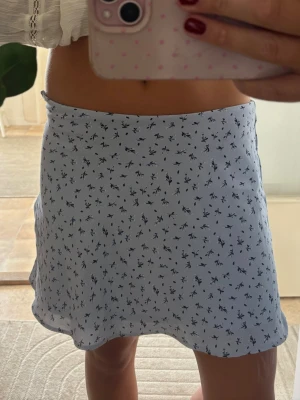 Ljusblå blommig kortkjol - Supersöt ljusblå kortkjol med små mörkblå blommor. Kjolen har en lätt utsvängd form och är gjord i ett tunt, mjukt material som känns perfekt för varmare dagar. Enkel att matcha med både toppar och stickade tröjor. Storlek 40 men liten i storlek