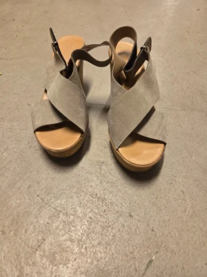 Beige sandaler i mocka - Säljer ett par snygga beige sandaler med kilklack och breda korslagda remmar i mocka. Sandalerna har öppen tå och justerbar rem runt hälen. Perfekta för dig som gillar stilrena och bekväma skor med lite höjd.
