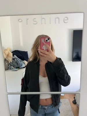 Svart bomberjacka Alpha Industries - Snygg svart bomberjacka från Alpha Industries med klassisk röd tag på ärmen och dragkedja framtill. Jackan har ribbade muddar vid ärmslut och midja samt fickor med tryckknappar. Tillverkad i glansigt nylonmaterial som ger en cool look.