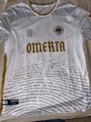 Vit och guld Omerta t-shirt två tröjor svart o vit - Säljer en vit t-shirt från Omerta med guldfärgade detaljer och tryck. Tröjan har rund halsringning, korta ärmar och ett unikt mönster i grått nedtill samt logga och text framtill. Perfekt för dig som gillar streetwear och snygga prints.