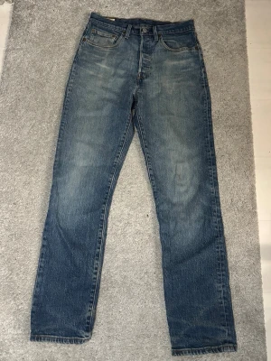 Levi's 501 blå - Klassiska blå jeans från Levi's med rak passform. Jeansen har en snygg tvättad look och är tillverkade i slitstark denim. Perfekta för en avslappnad stil och passar till det mesta. Levi's patch bak i midjan och diskreta sömmar. W27 L30 (lite slitna nertill)