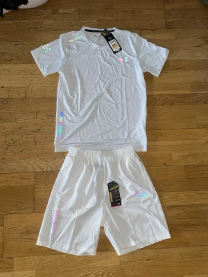 Vitt träningsset från under armour (reflex) - Säljer ett fräscht vitt träningsset från under armour med t-shirt och shorts. Setet har holografiska detaljer och loggor på både tröja och shorts. Materialet är lätt och andas, perfekt för träning eller sportaktiviteter. Elastisk midja på shortsen för skön passform. Det står storlek M men passar både S och M.