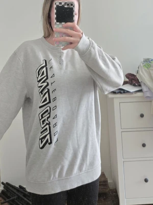 Grå Santa Cruz sweatshirt - Säljer en grå sweatshirt från Santa Cruz med stort svart tryck längs framsidan där det står 'Santa Cruz Skateboards'. Tröjan har rund halsringning och ribbade muddar vid ärmslut och nederkant. Perfekt för dig som gillar streetwear och skatekultur.