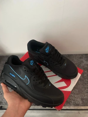 Nike Air Max 90 svart/blå sneakers - Nike Air Max 90 sneakers i svart med blå detaljer och logga. Skorna har en klassisk siluett med synlig Air Max-dämpning i sulan, mesh och läderpaneler samt blå Swoosh och branding. Perfekta för dig som gillar streetwear och sportig stil.