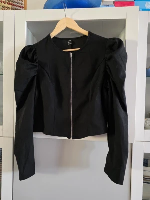 Svart croppad jacka från SHEIN - Trendig svart jacka från SHEIN med croppad passform och puffiga axlar. Jackan har lång ärm och dragkedja framtill. Perfekt för dig som vill ha en edgy look med extra attityd.