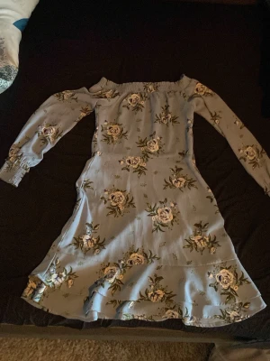 Blommig ljusblå långärmad klänning - Söt ljusblå klänning med blommigt mönster och långa ärmar. Klänningen har offshoulder-design med resår upptill och vid ärmslut. Dragkedja på sidan så att man lättare får på den! Volangdetalj nertill ger extra flow. Perfekt för vår och sommar!  