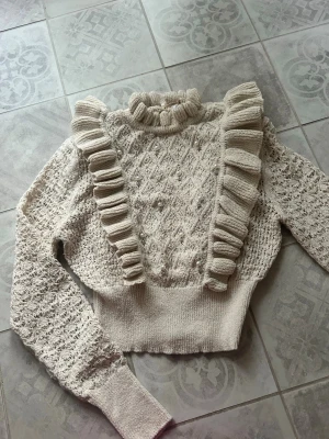 Beige stickad tröja med volanger - Supersöt beige stickad tröja med höghalsad krage och volangdetaljer framtill. Tröjan har ett fint strukturmönster och långa ärmar med spetsstickning. Perfekt för dig som gillar romantisk och trendig stil.